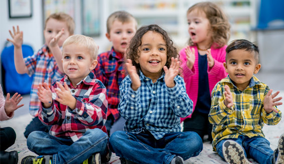 Children sit in 2 rows on a carpet while clapping