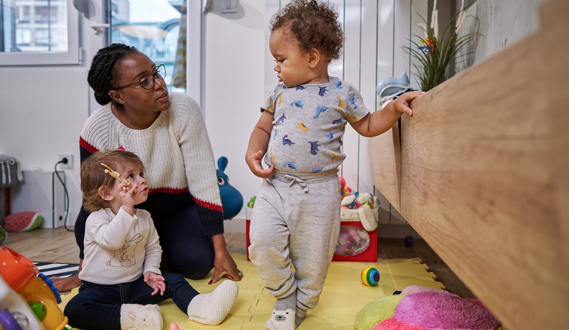 A day care provider sits with a toddler as 1 stands holding on