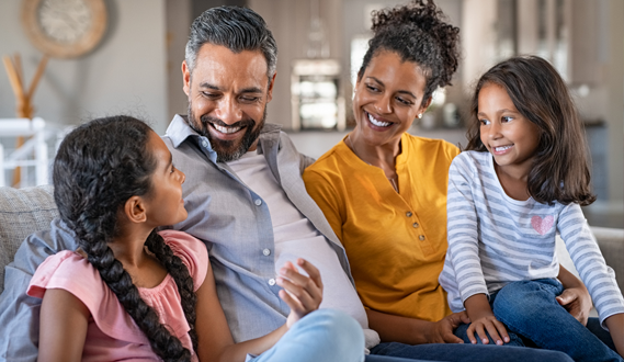 A family of four sits on the couch smiling and talking.