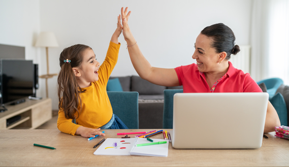 A woman sitting at a computer high fives a young girl that has a workbook in front of her.