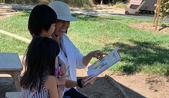 A preschool teacher sits with 2 students on an outside bench reading a book.