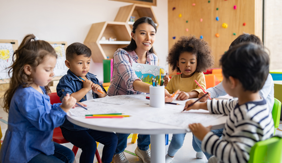 A smiling teacher engages with a diverse group of students as they draw.