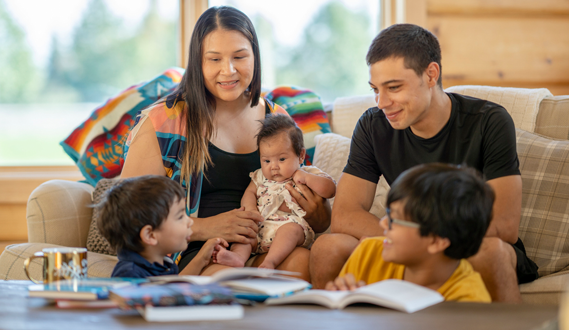 A native family sites on a couch while the dad reads to their 3 young children.
