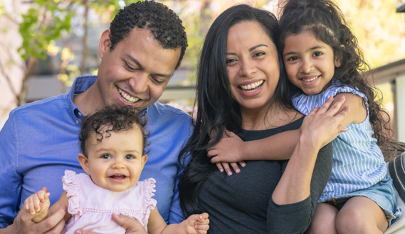 A smiling Hispanic family with 2 young daughters pose for a picture.