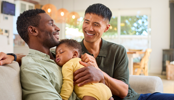 A smiling gay couple holds their sleeping baby.
