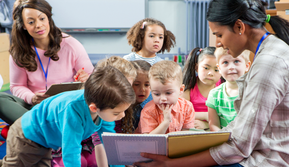 A teacher reads a book to a preschool class while another adult observes.