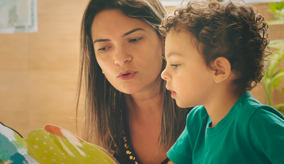 A woman sounds out words to a young boy as they read a book.