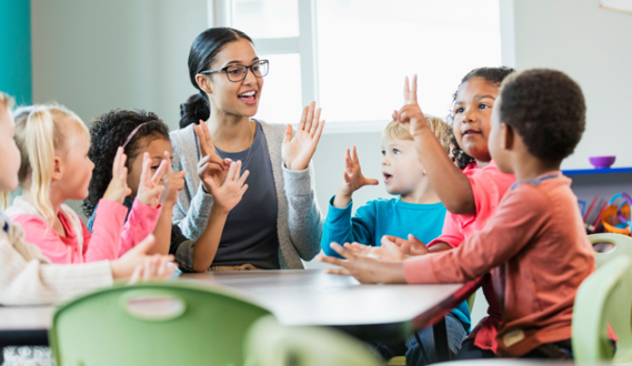 A teacher and her preschool age students hold up their hands and sing.