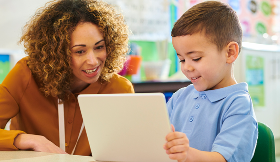 A smiling teacher helps a student working on a tablet.