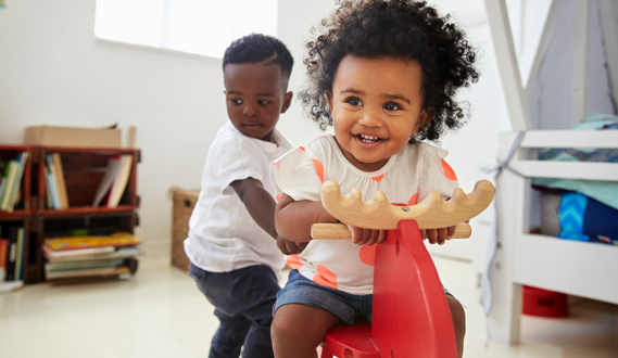 A toddler girl sits on a ride-on toy while a boy plays behind her.