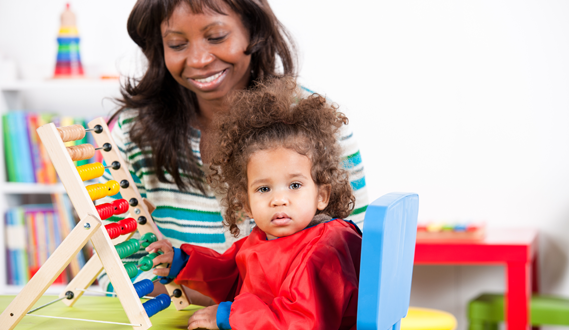 A daycare provider sits with a toddler playing on an abacus.