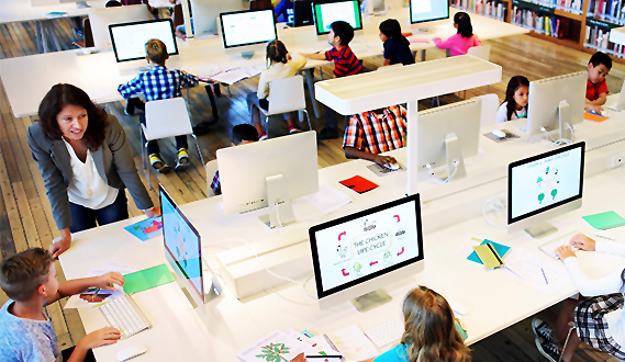 A teacher looks over a student's shoulder in a computer lab while other students work.
