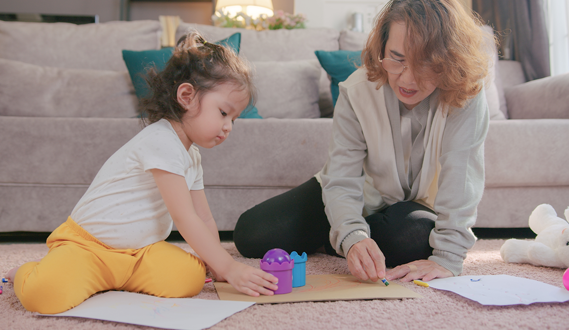 An older child care provider colors with a preschool-aged child on her living room floor.