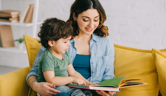 A woman sits on a yellow couch with a toddler boy on her lap reading a book.
