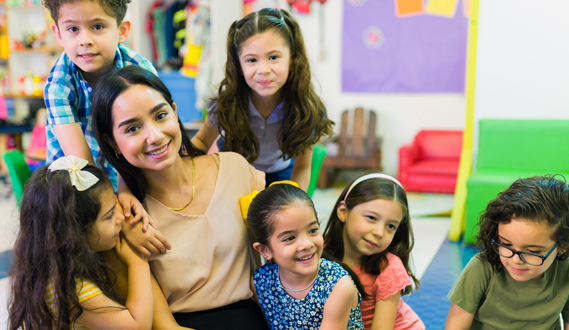 A Hispanic teacher sits with her smiling DLL students.