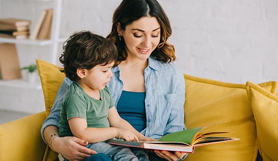 A woman reads a boo to a young preschooler on a yellow couch.