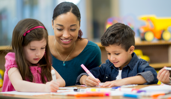 A smiling teacher sits behind 2 children working on an assignment.