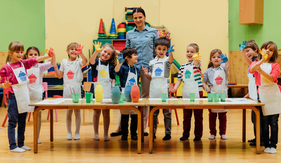 A teacher stands with students in aprons while fingerpainting.