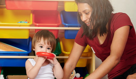 A woman sits with a young toddler chewing on a plastic ring.