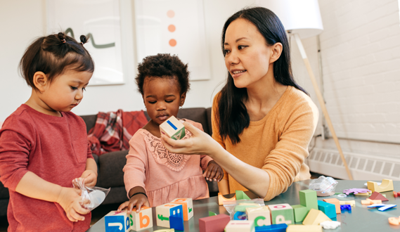 A teacher holds up a square wooden block to her two preschool students.