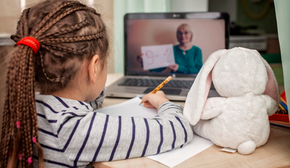 A young child sits with her stuffed animal and watches a virtual lesson on a laptop.
