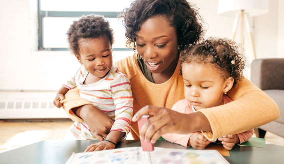 A sitting caregiver holds a toddler in each arm while she reads a book to them.