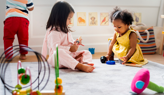 Two toddlers play with stacking rings on a carpet.