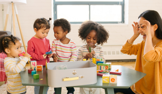 A teacher holds her hands by her eyes as if she's waiting to see a surprise, while her young preschoolers are gathered around an activity in a box.