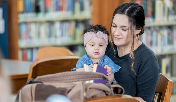 A woman holds a toddler and reads to her at a library table