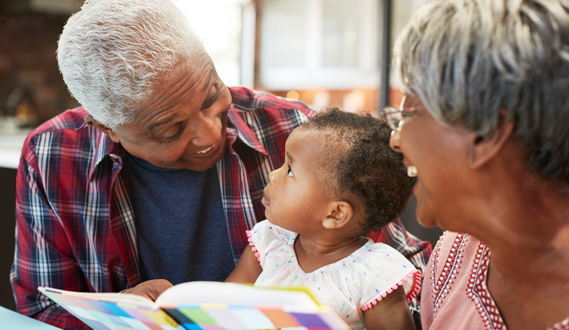 An elderly couple reads a book to an infant girl.
