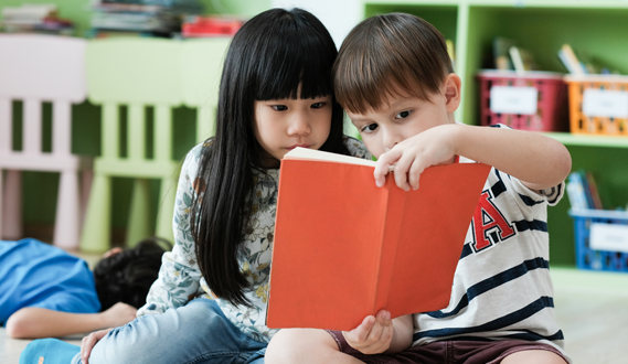 A young boy holds up a book as he reads it with a girl.