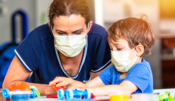A daycare provider and a young boy play togeter wearing masks.