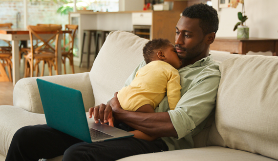 A father holds his sleeping baby as he uses a laptop