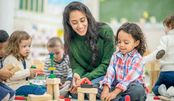 A preschool teacher sits with a young boy building with blocks.