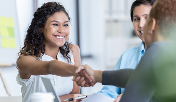 A smiling woman shakes hands with someone over a table.