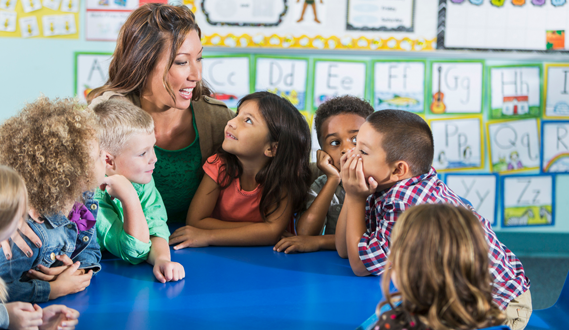 A teacher sits at a round table with her students as one girl beams up at her.