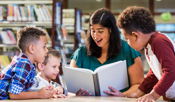 A woman reads to 3 young boys at a table in the library.