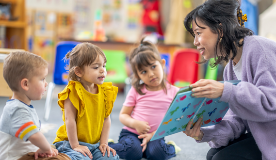 A teacher holds up a book she is reading to 3 young students.
