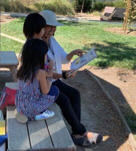 A preschool teacher reads with two students on an outdoor bench.