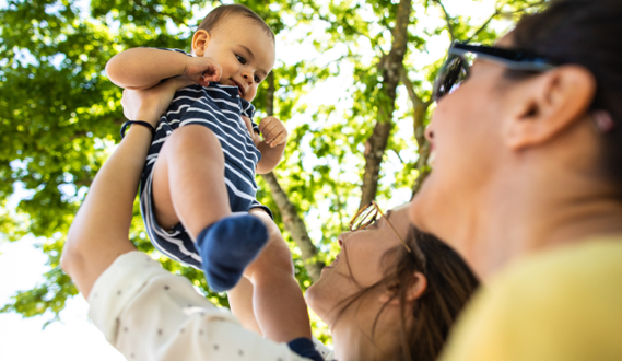 Smiling parents hold up a baby with trees in the background.