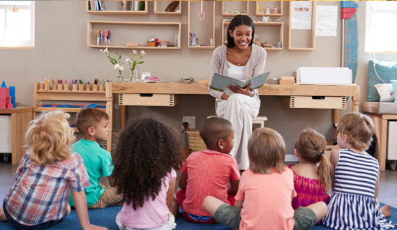 A teacher sits in a chair reading a book to her class as they sit on a carpet.