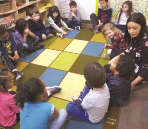 A teacher sits in a circle with young students in a classrrom.