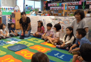 A teacher kneels on a classroom carpet with her students during circle time.