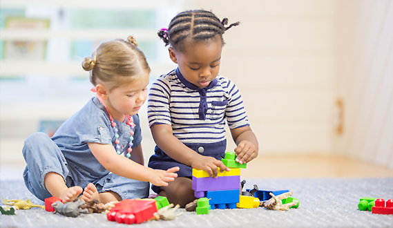 Two toddler girls, with their hair in 2 buns, play together on a carpet.