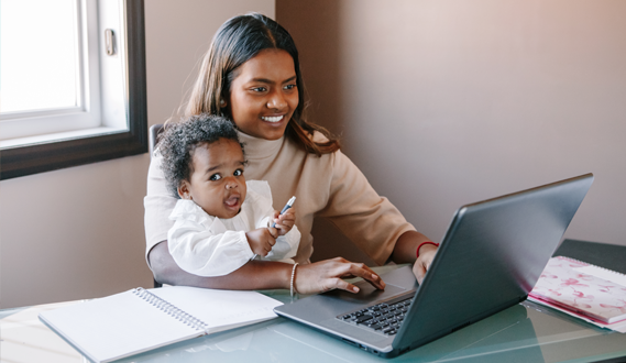 A mother works on a computer while holding her baby.