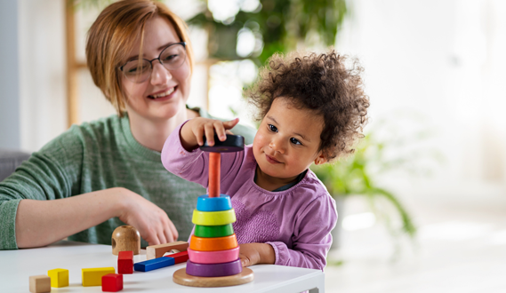 An in-home child care provider smiles as a toddler stacks toy rings.