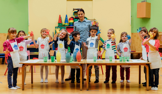 A teacher poses with her art students who are wearing aprons and painting.