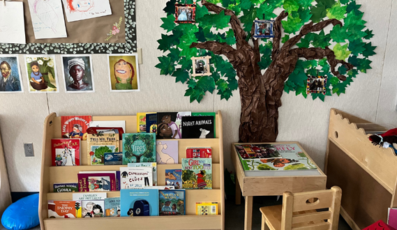 A library corner in a preschool classroom that has a tree panted on the wall.