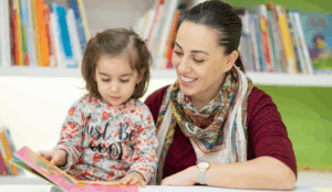 A teacher sits with a young girl that is learning to read in a school library.