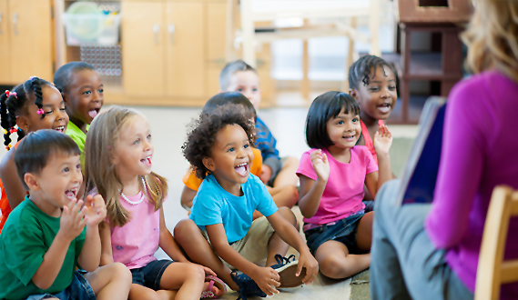 CHildren sit in 2 rows on a classroom carpet smilng and laughing at a book the teacher is reading.